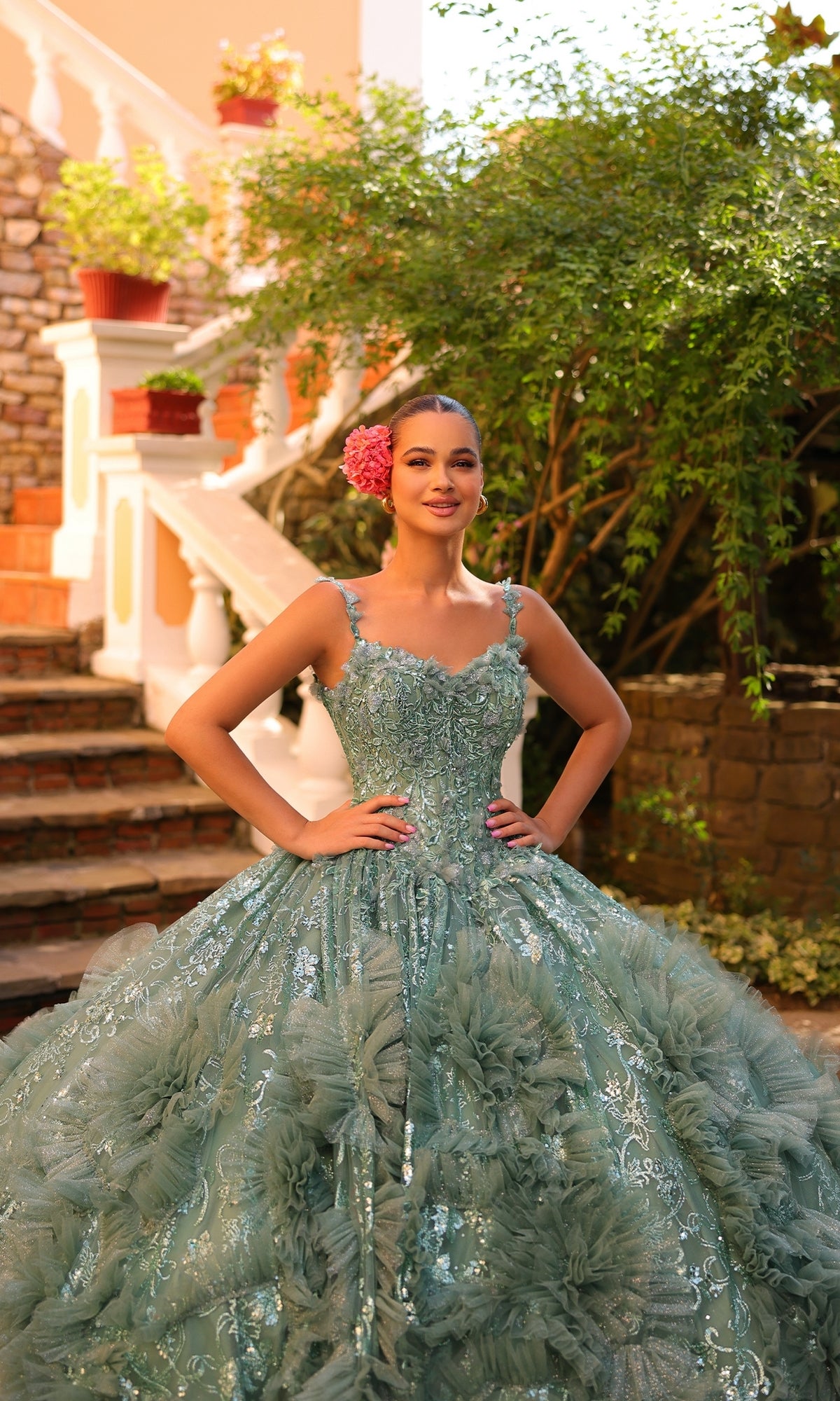 A young woman poses outdoors in the Quinceanera Dress 54314 By Amarra, a light blue-green ball gown featuring floral accents, a sheer cape train, and a pink flower in her hair, with her hands on her hips.